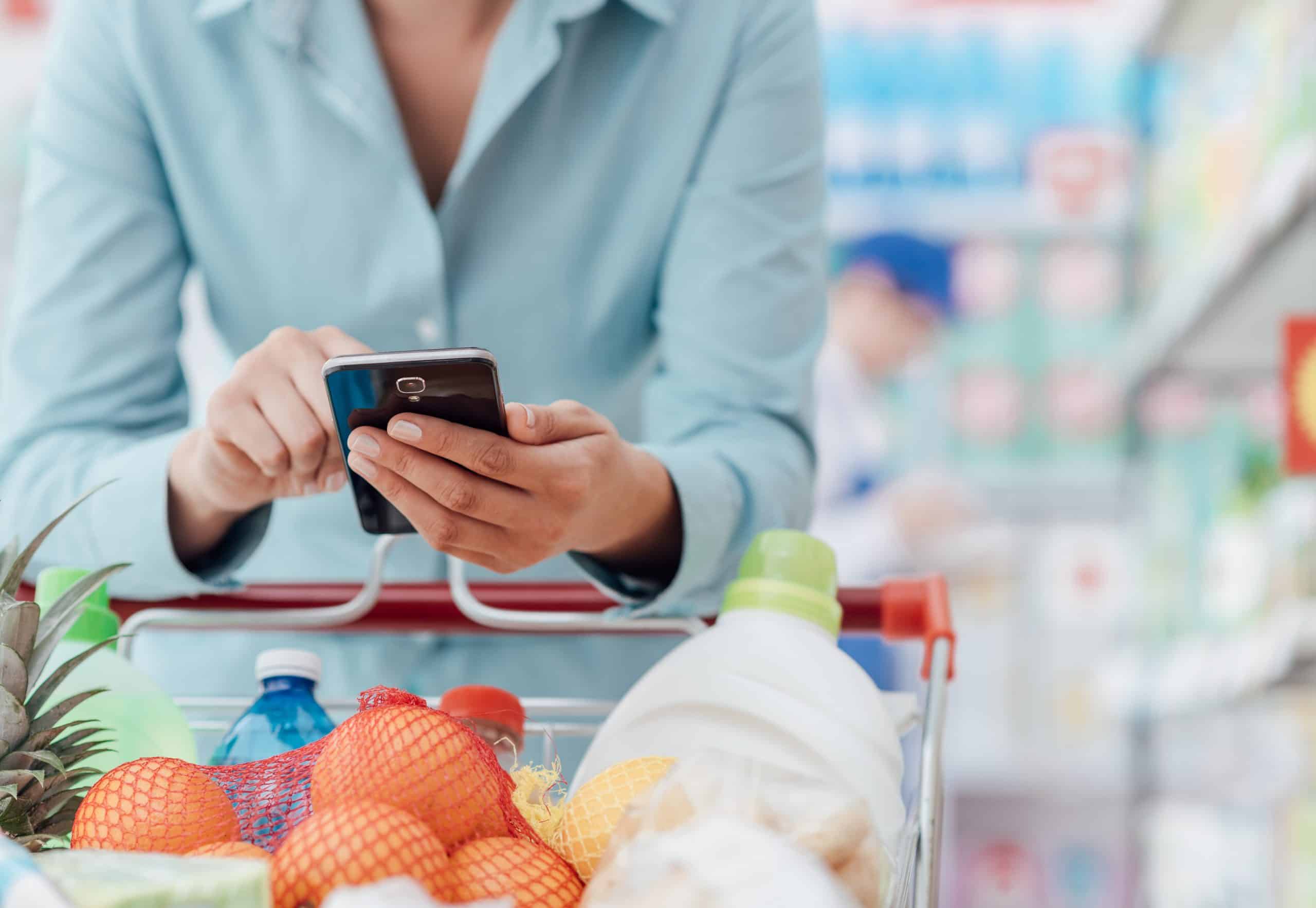 More frugal than your spouse - Woman in blue button-down shirt looking at phone app by her supermarket shopping cart